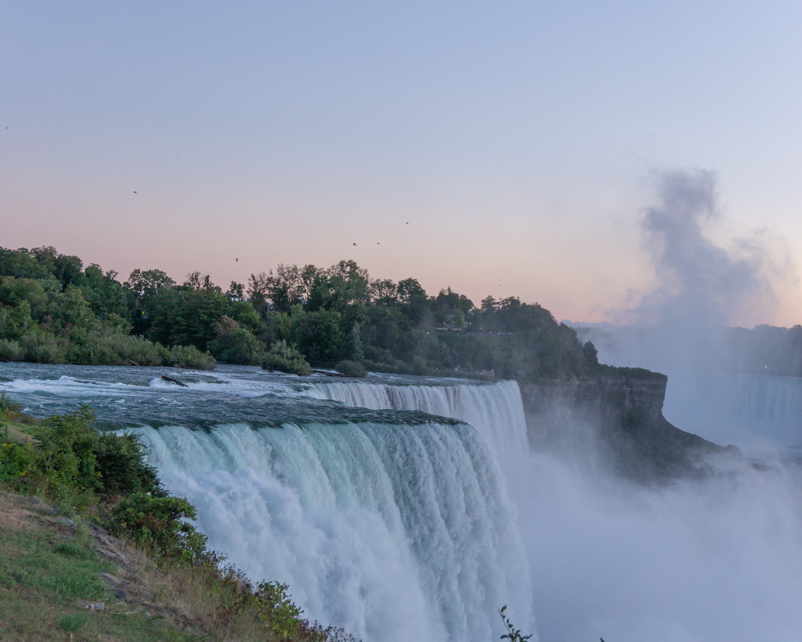 The American Falls at sunrise under a soft pink sky, viewed from the New York side